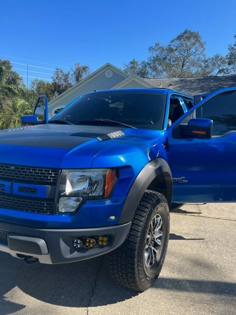 Blue Ford Raptor truck with tinted windows and open doors parked outside a house on a sunny day.