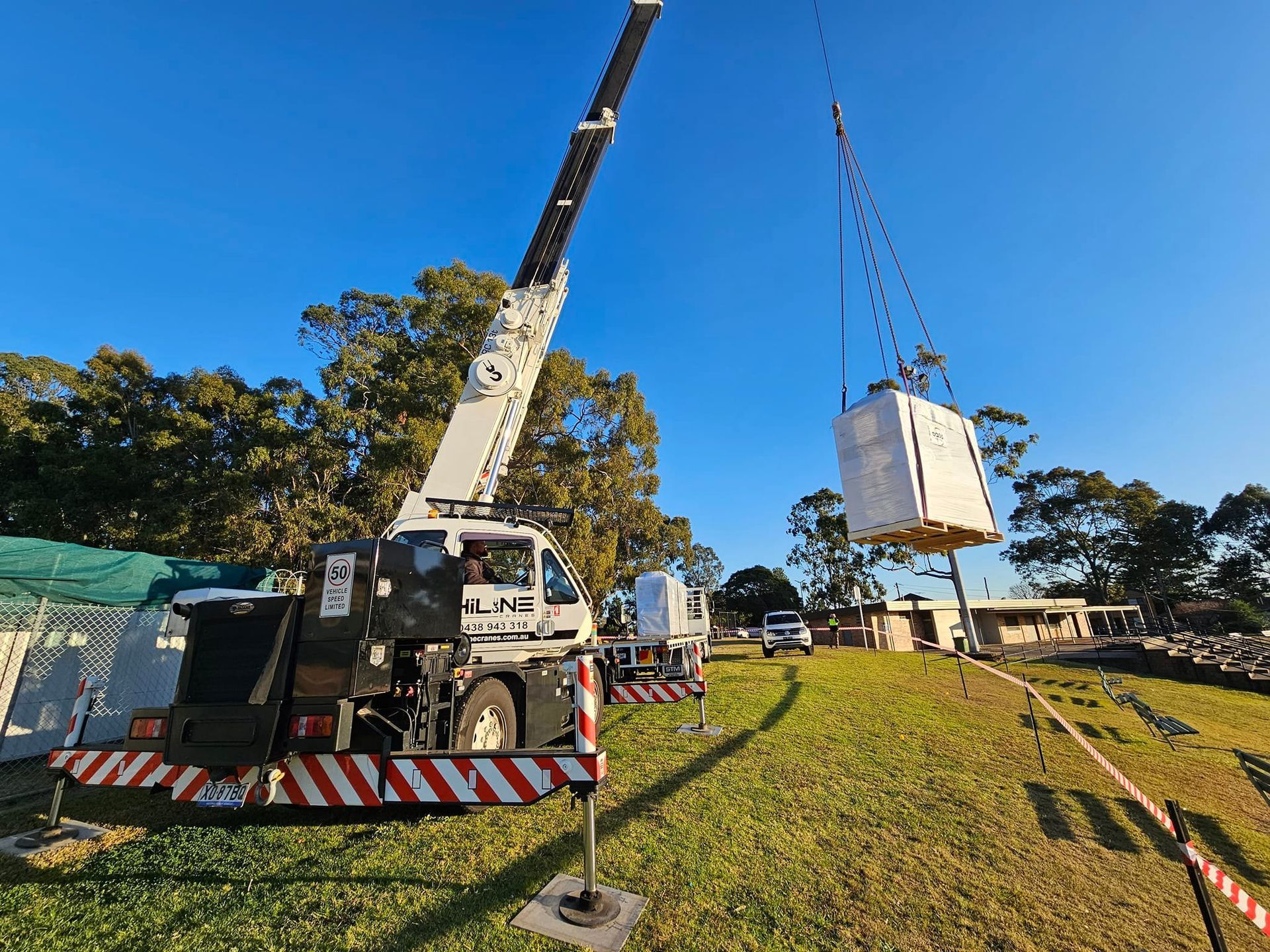 A HiLine crane lifting a large white box on a grassy field, sunny day.