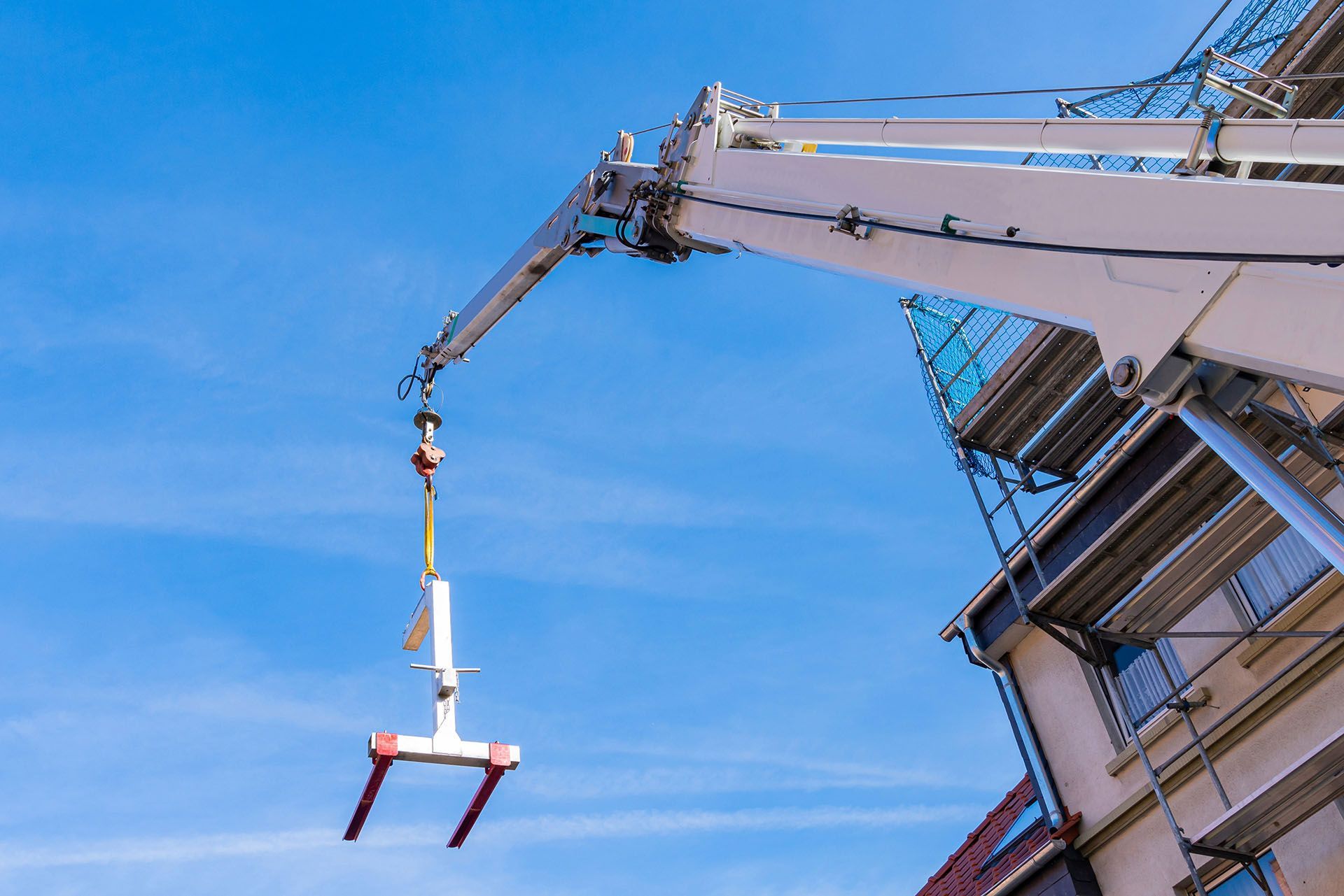 A crane lifting a white construction tool against a bright blue sky, next to a building.