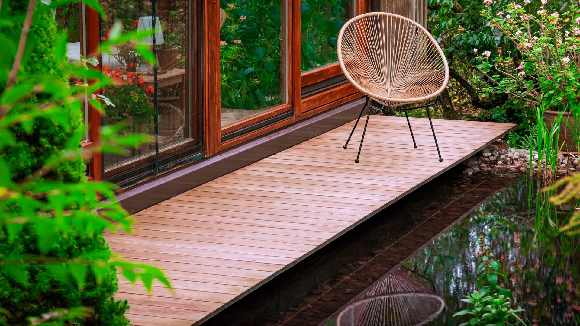 Woven chair on a wooden deck by a pond, with a glass door in the background and lush greenery.
