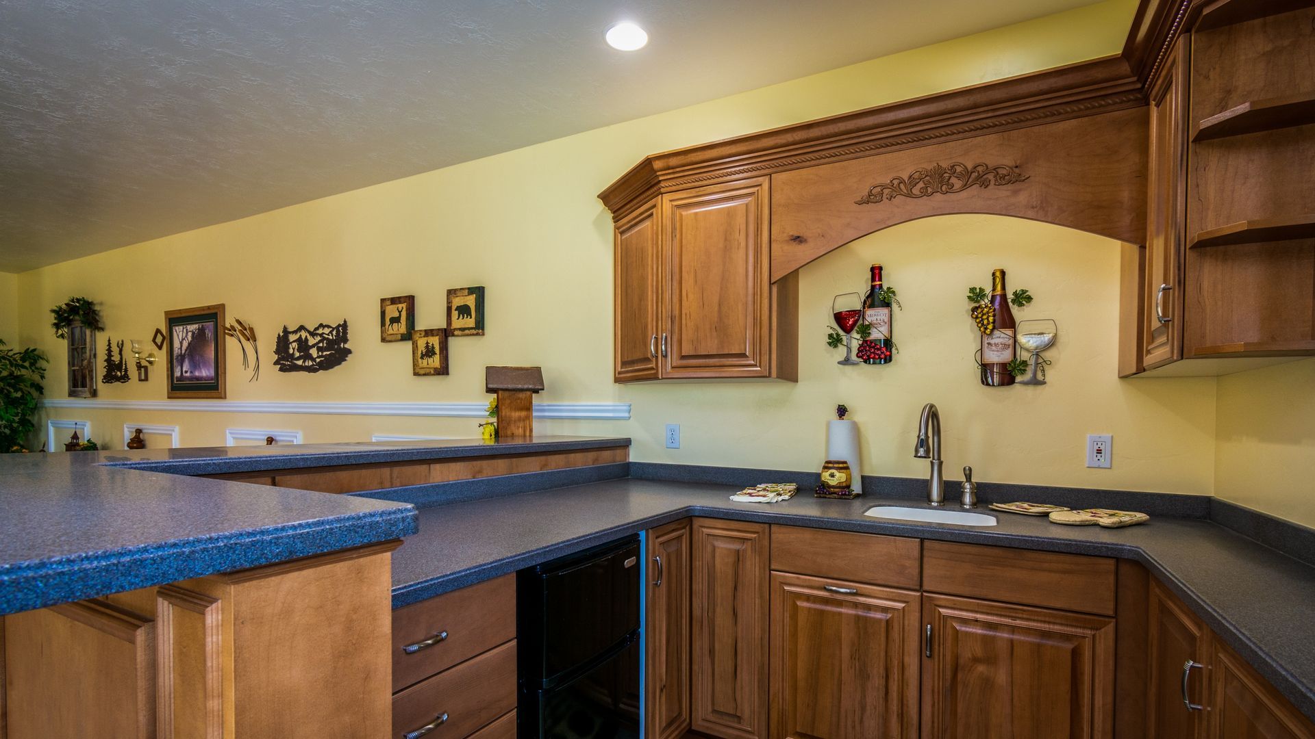 L-shaped kitchen with brown cabinets, blue countertops, and yellow walls. Decorative wine racks hang above the sink.