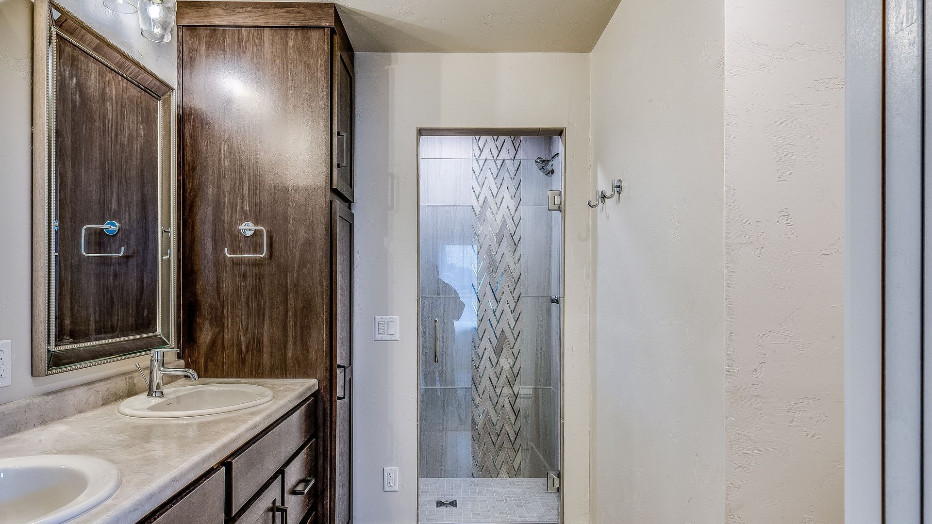 Bathroom with a double vanity, tall storage cabinet, and walk-in shower with decorative glass panel.