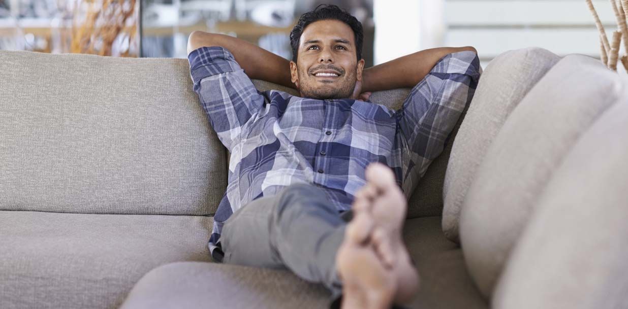 A man is sitting on a couch with his feet up and his hands behind his head.