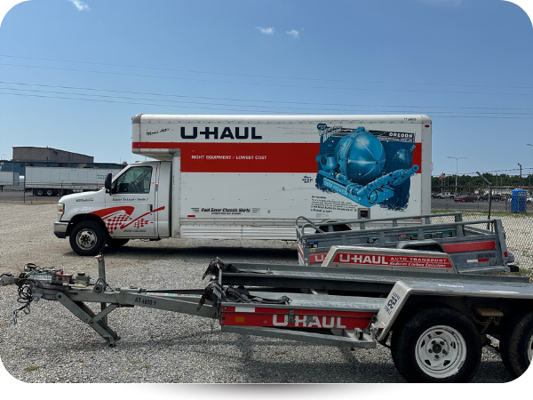 A white U-Haul moving truck parked on a gravel lot with a U-Haul vehicle transport trailer in the foreground.