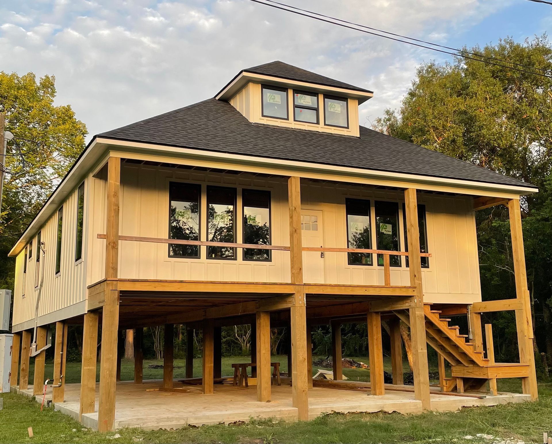 A light-colored two-story house on stilts. It has a porch and a black roof, with windows and a staircase.