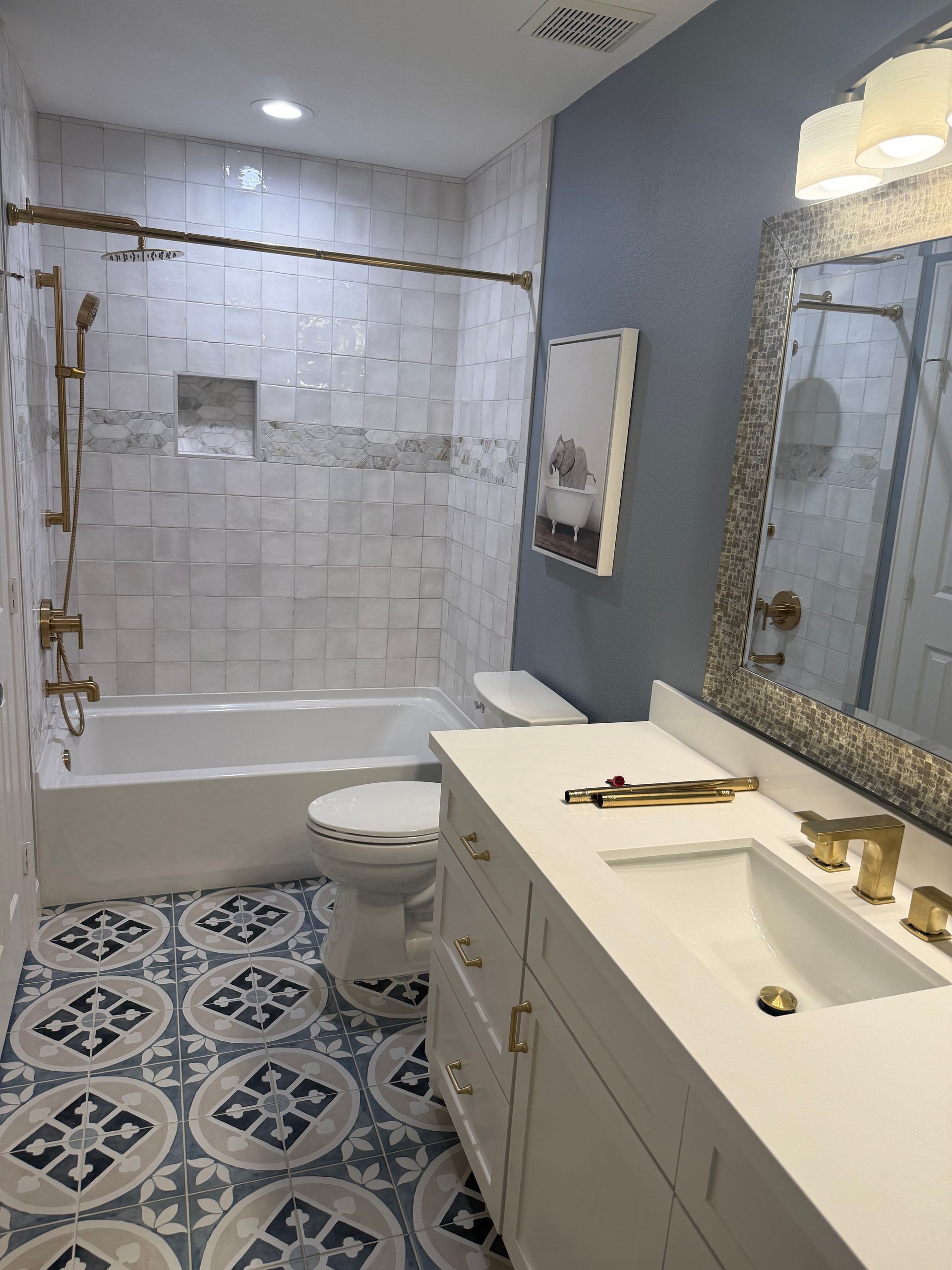 Bathroom with blue patterned floor tiles, white vanity, and blue wall. Gold fixtures and a framed picture add detail.