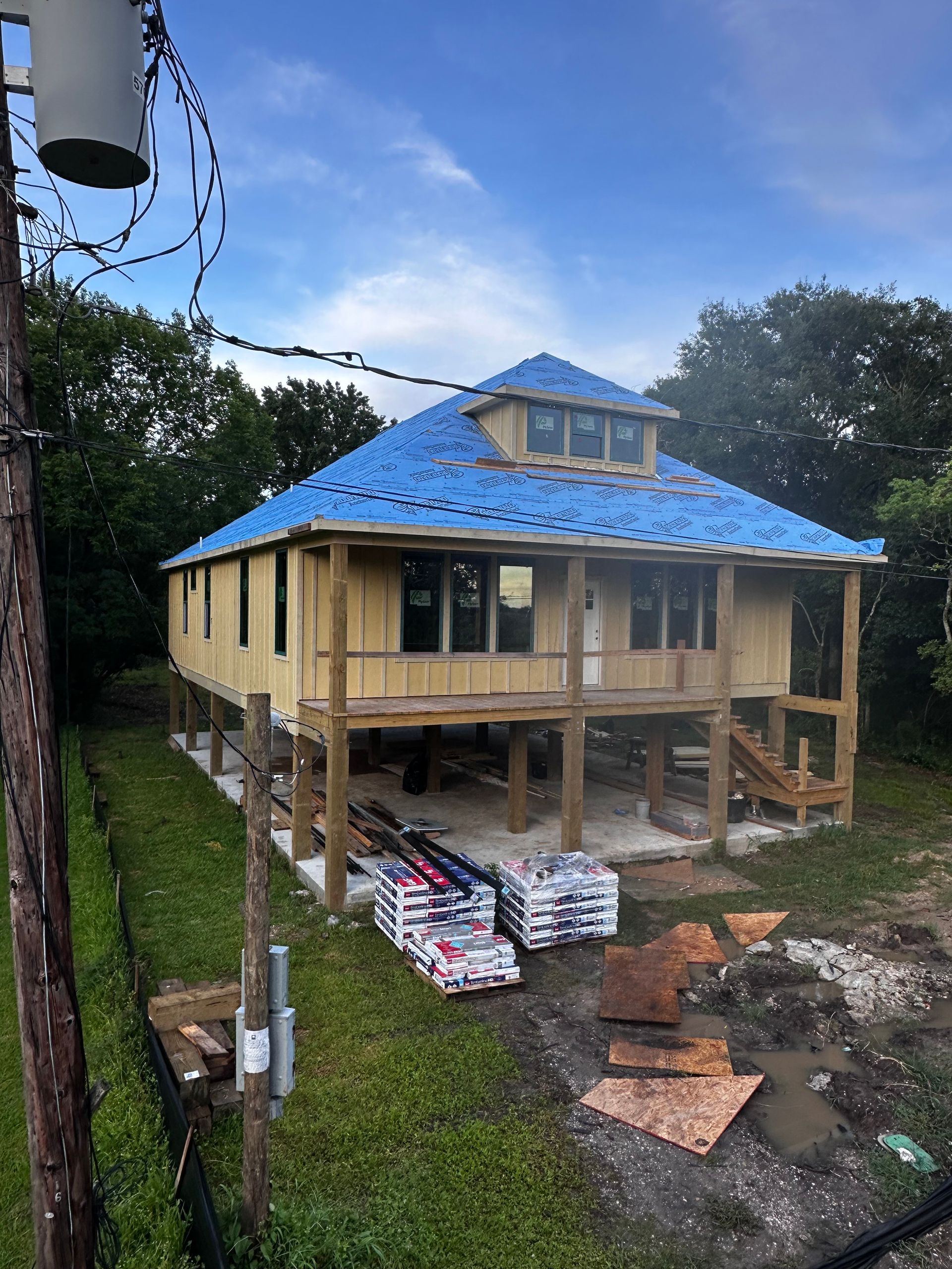 House under construction, raised on wooden pilings with blue roofing. Materials are stacked in front on the green lawn.