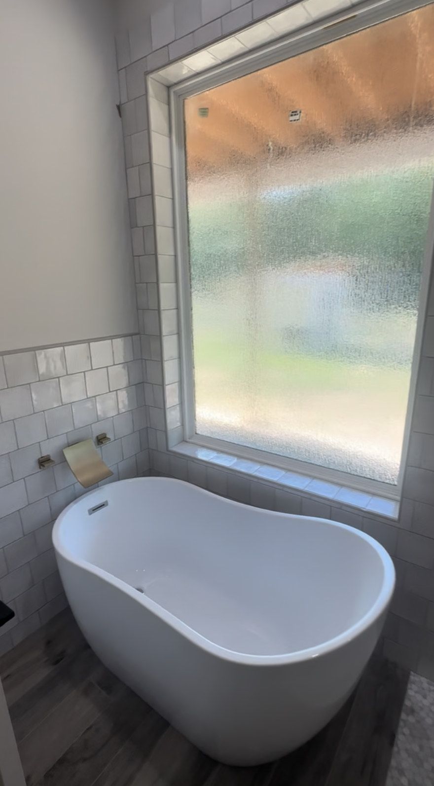 A modern white bathtub sits beside a window in a bathroom. The window has frosted glass and provides a view of green foliage.