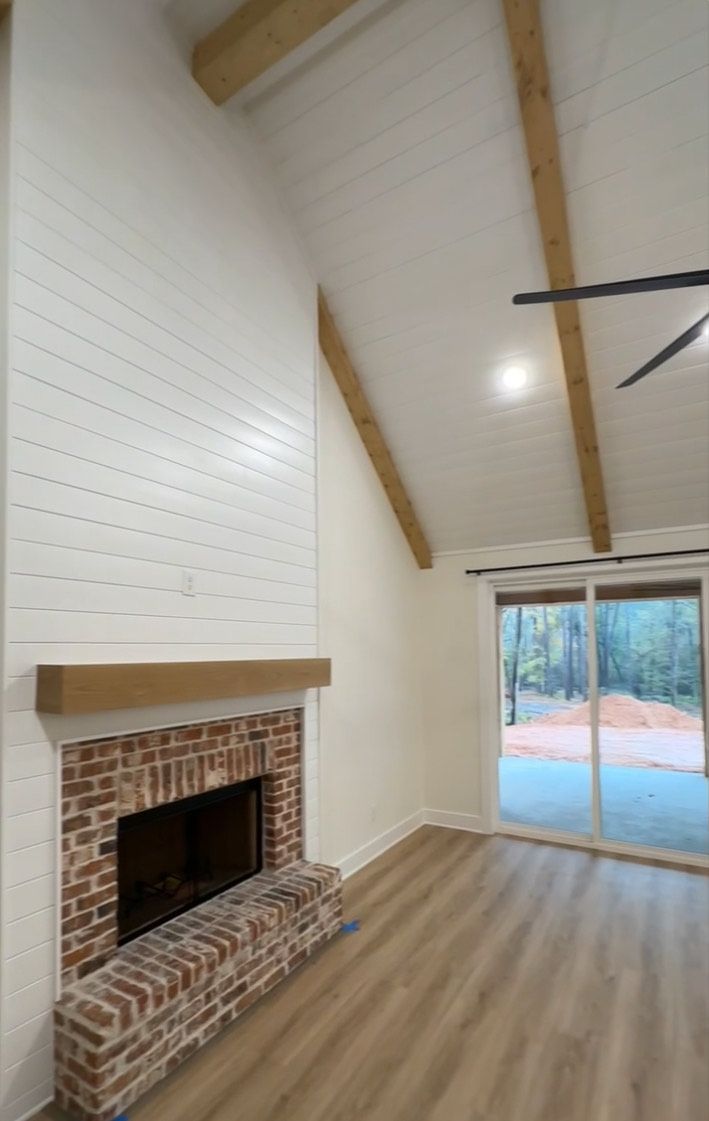 Living room with brick fireplace, white shiplap walls, exposed wood beams, and a sliding glass door overlooking a patio.
