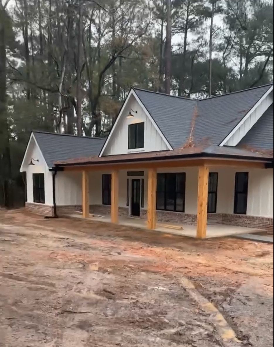 White farmhouse with a dark gray roof and a wooden porch under construction in a wooded setting.