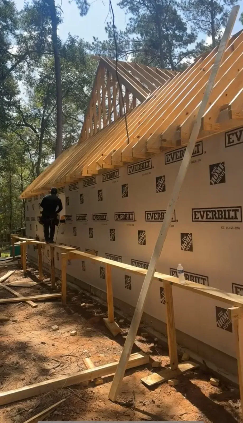 Construction worker on a roof, installing trusses. Building's exterior wall is covered in white weather-resistant barrier.