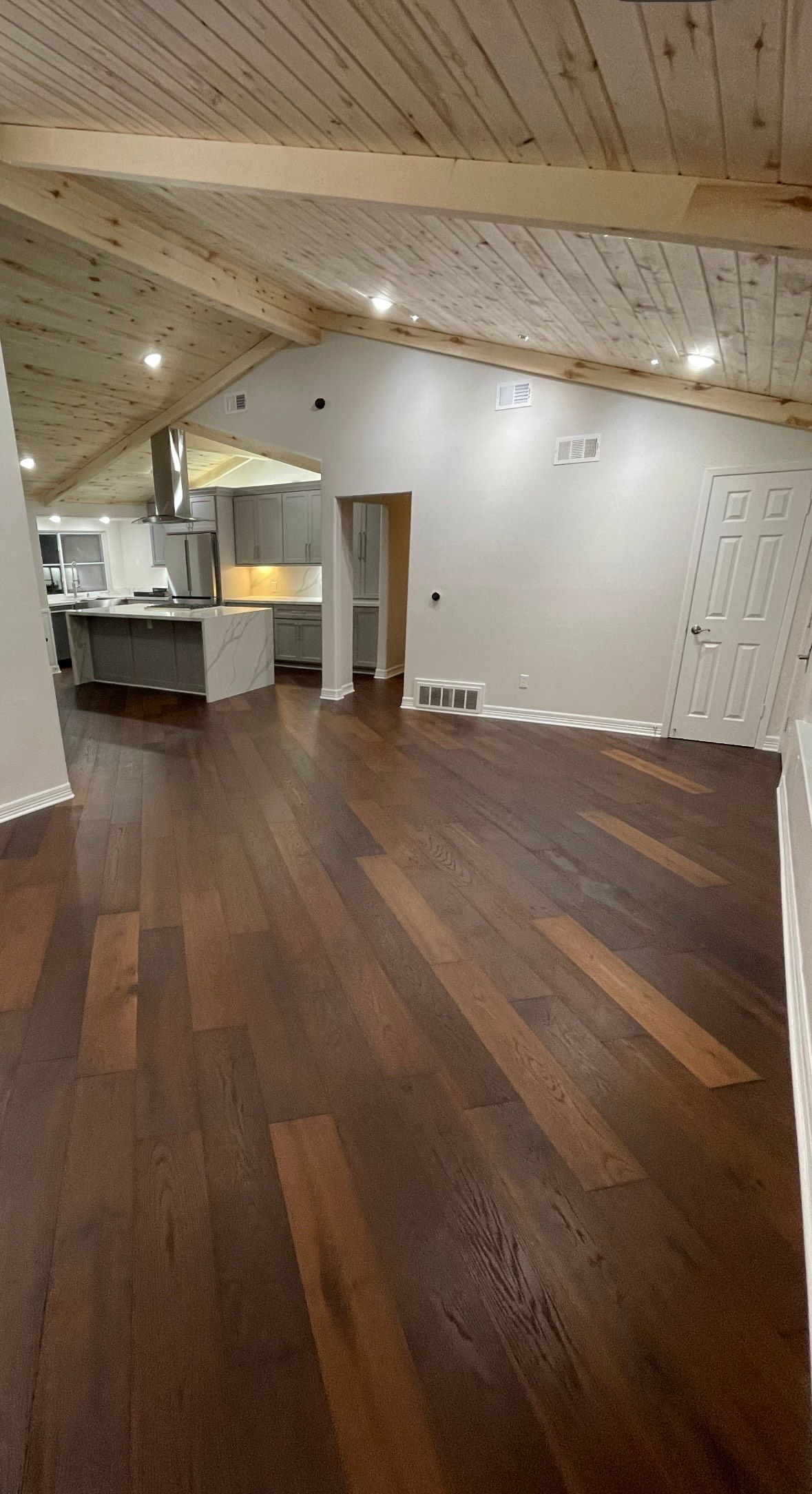 Wooden floors in a room with an angled, wood-beam ceiling. A kitchen with grey cabinets is visible in the background.