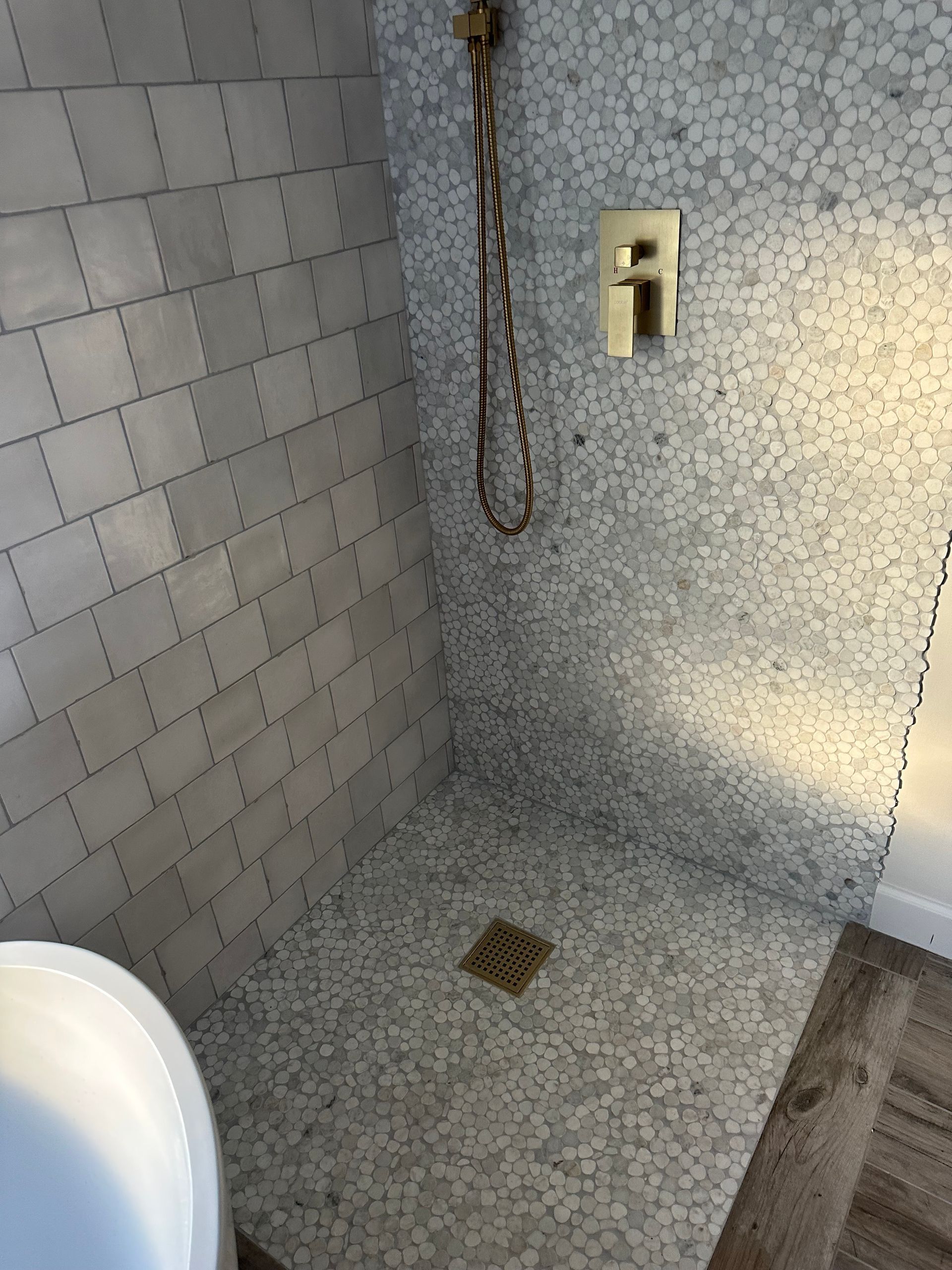 Shower with gray pebble tile floor and walls, gold fixtures, and square tile on one wall.