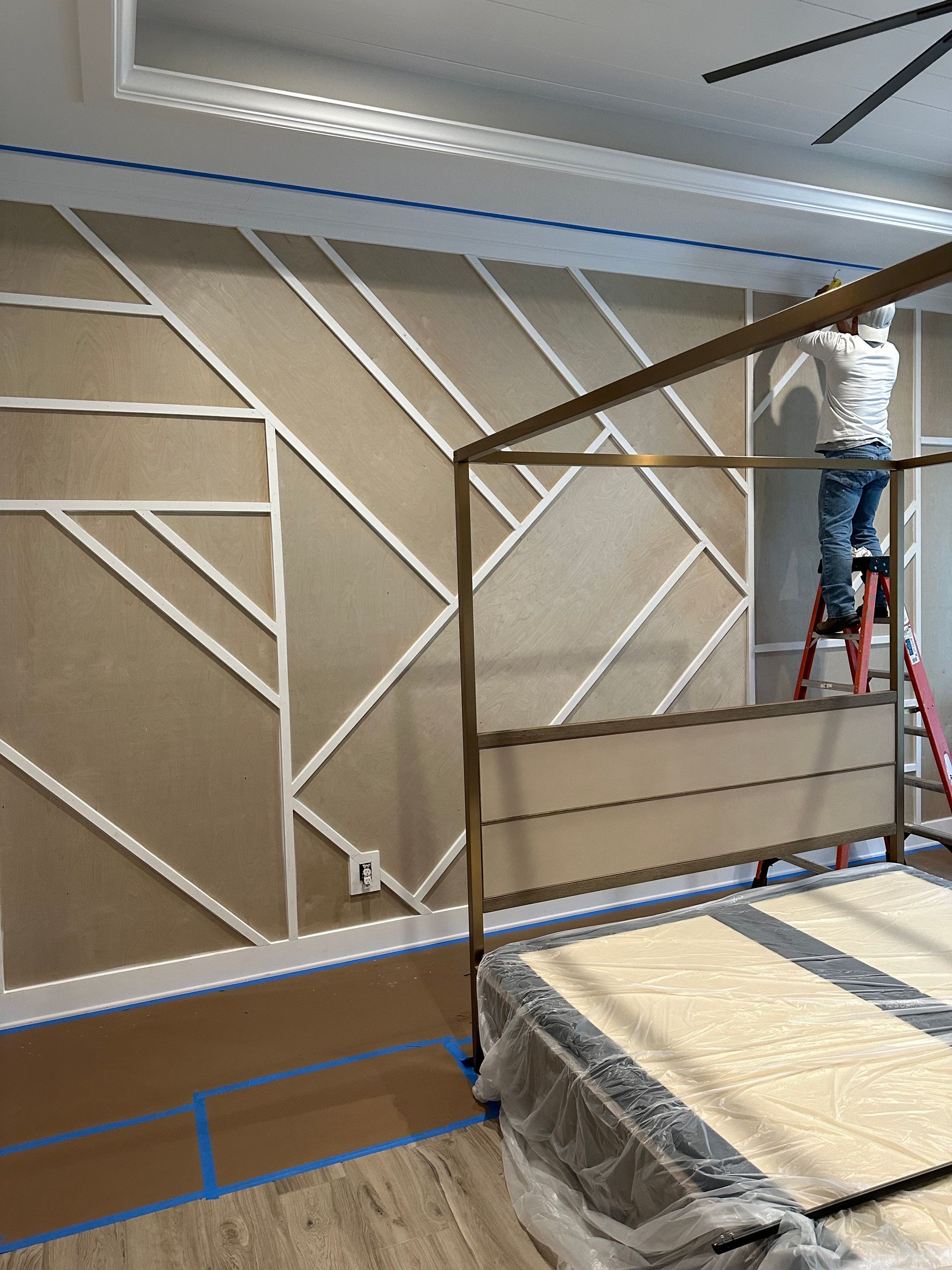 Person on ladder installing trim on a geometric accent wall in a bedroom. A four-poster bed frame is in the foreground.