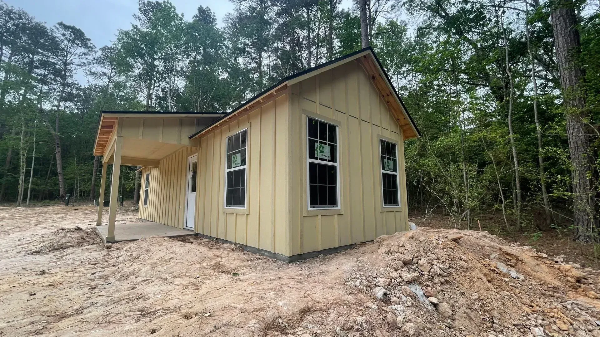 Small, light yellow house under construction, with porch, two windows, and dark window frames, surrounded by dirt and trees.