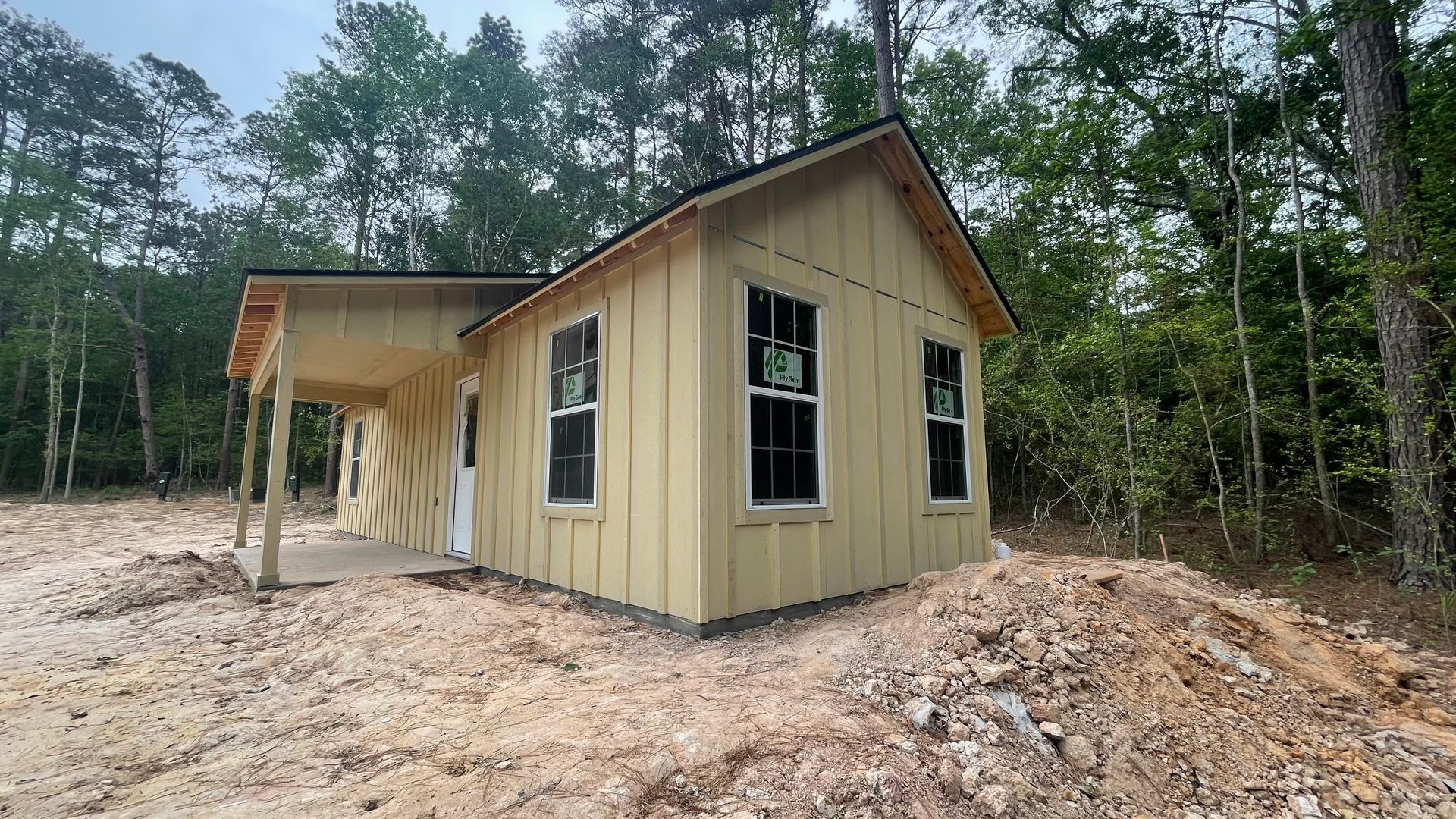 Small, yellow house under construction with a porch, surrounded by dirt and trees. Windows have black frames.