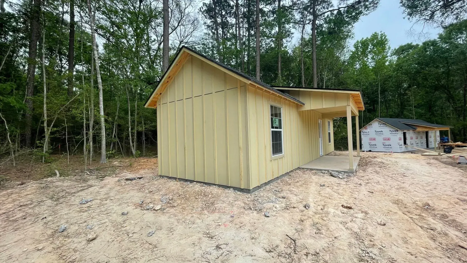A partially built, yellow-sided house with a porch sits in a sandy area surrounded by trees.