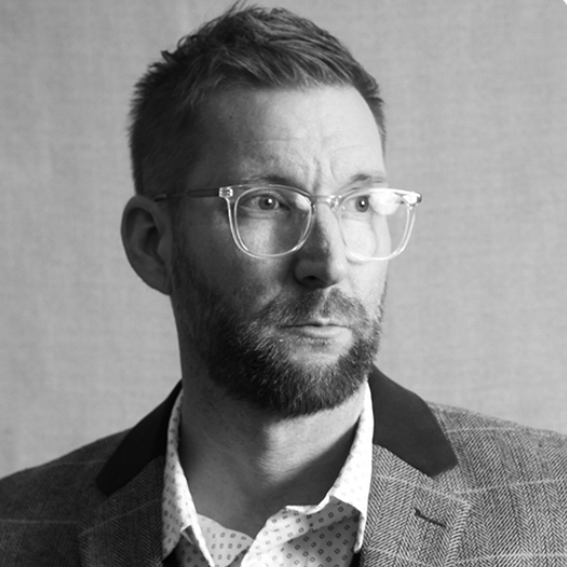 
A man with short hair, a beard, and clear-framed glasses, wearing a patterned shirt and a blazer with dark lapels, looking slightly to the side against a neutral background.
