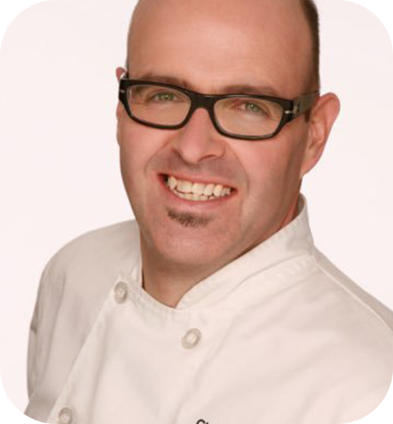 
A smiling man with glasses and a small goatee, wearing a white chef's jacket, facing slightly to the side against a light background.