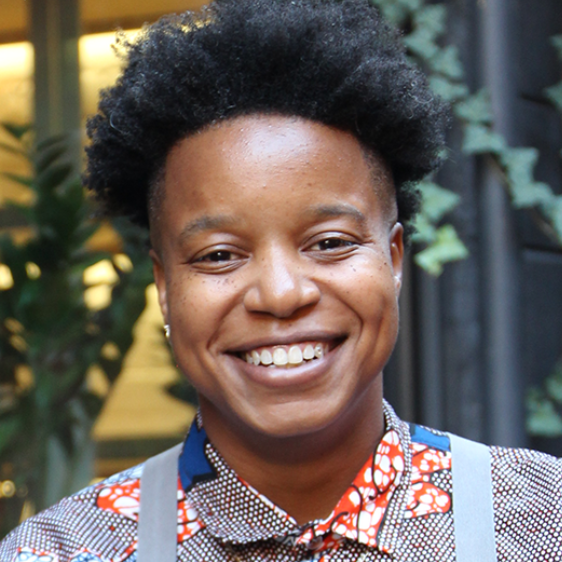 A smiling woman with short, curly black hair, wearing a colorful patterned shirt with suspenders, standing in front of a blurred indoor background.

