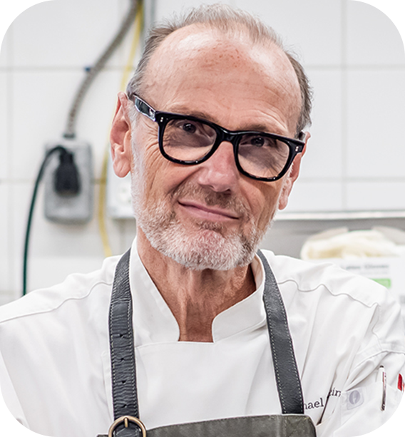 
Portrait of a slightly smiling man with a grey beard, wearing black glasses, a white chef's coat, and a dark apron, in a professional kitchen setting.

