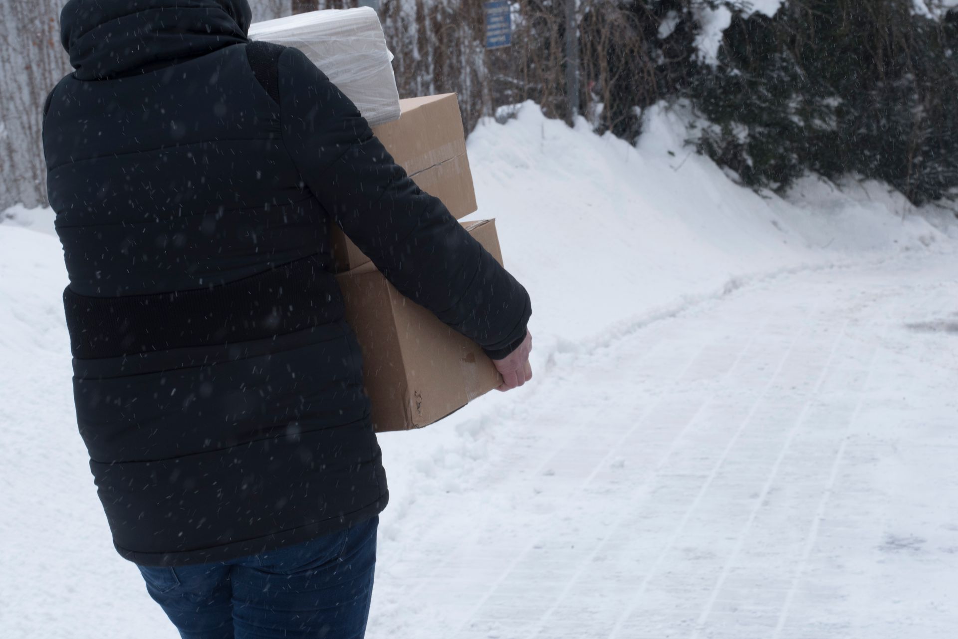 Person bundled in a black coat carries boxes and paper towels in a snowy outdoor setting.