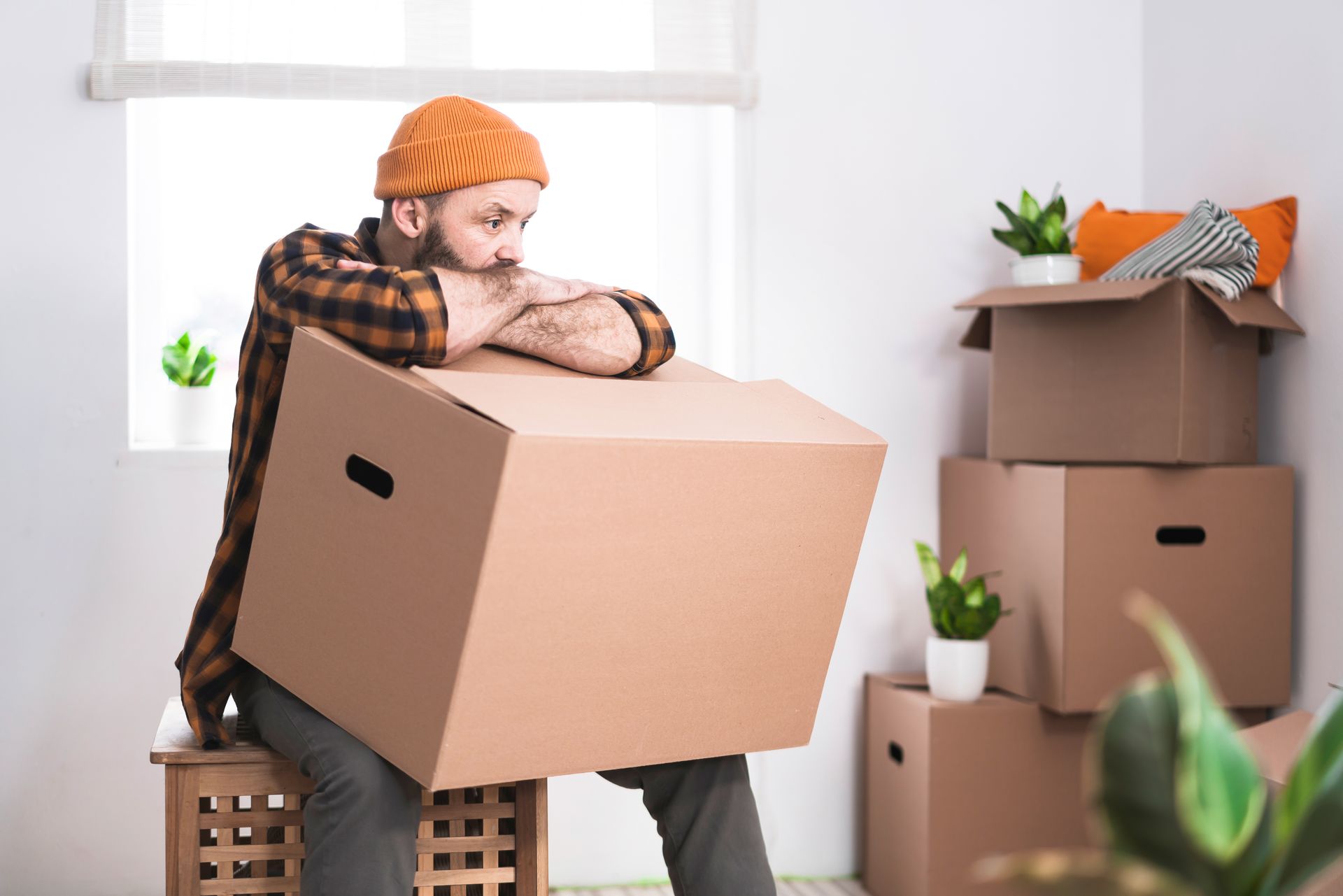 Man in orange beanie resting on a cardboard box, looking weary, surrounded by moving boxes in a room.