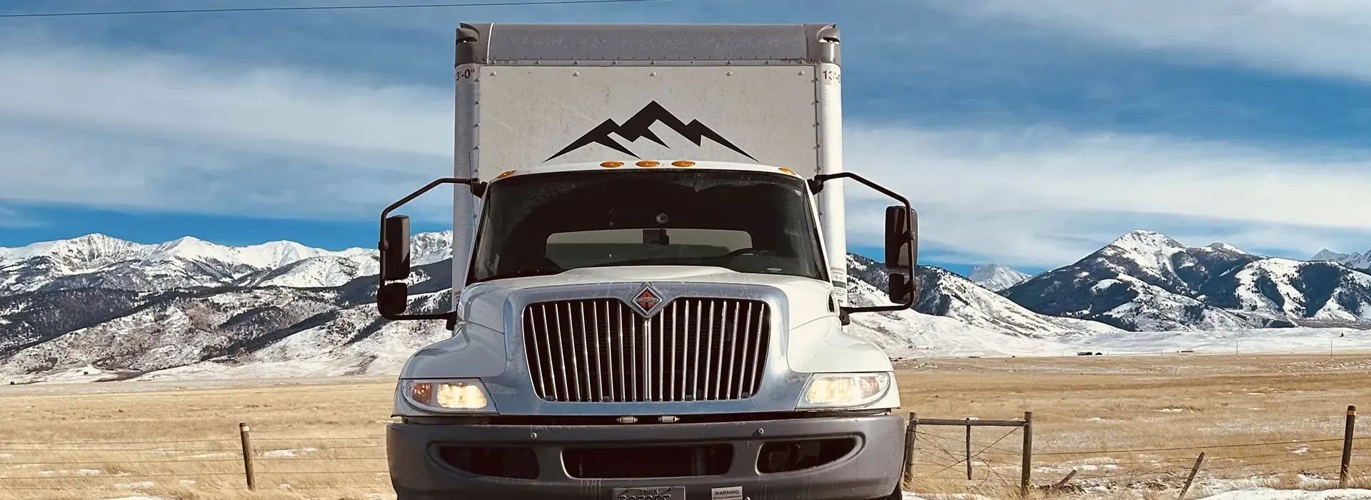 White box truck driving toward the viewer, with mountains and blue sky in the background.