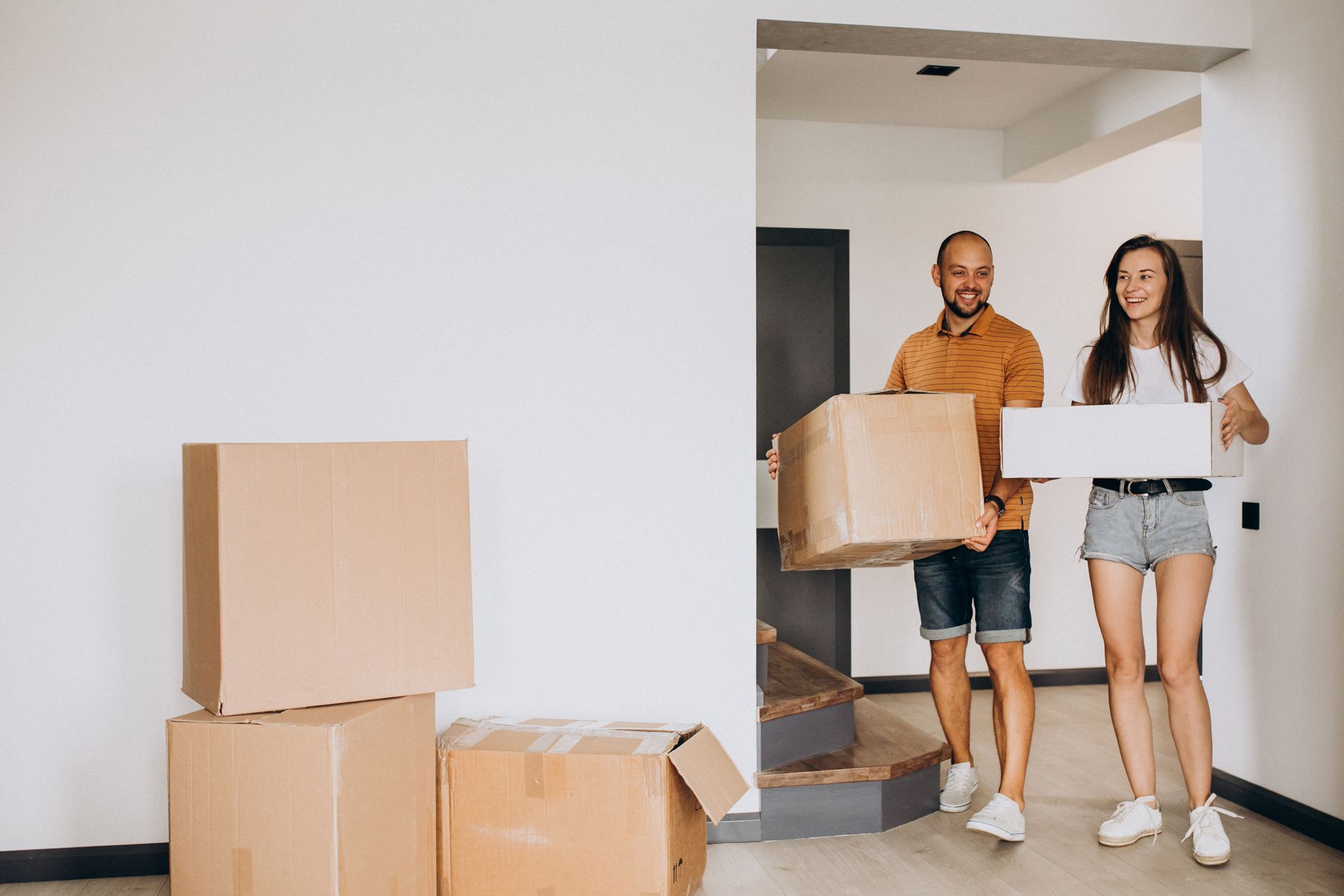 Couple carrying boxes into a new home; smiling, near a staircase and stacked boxes.