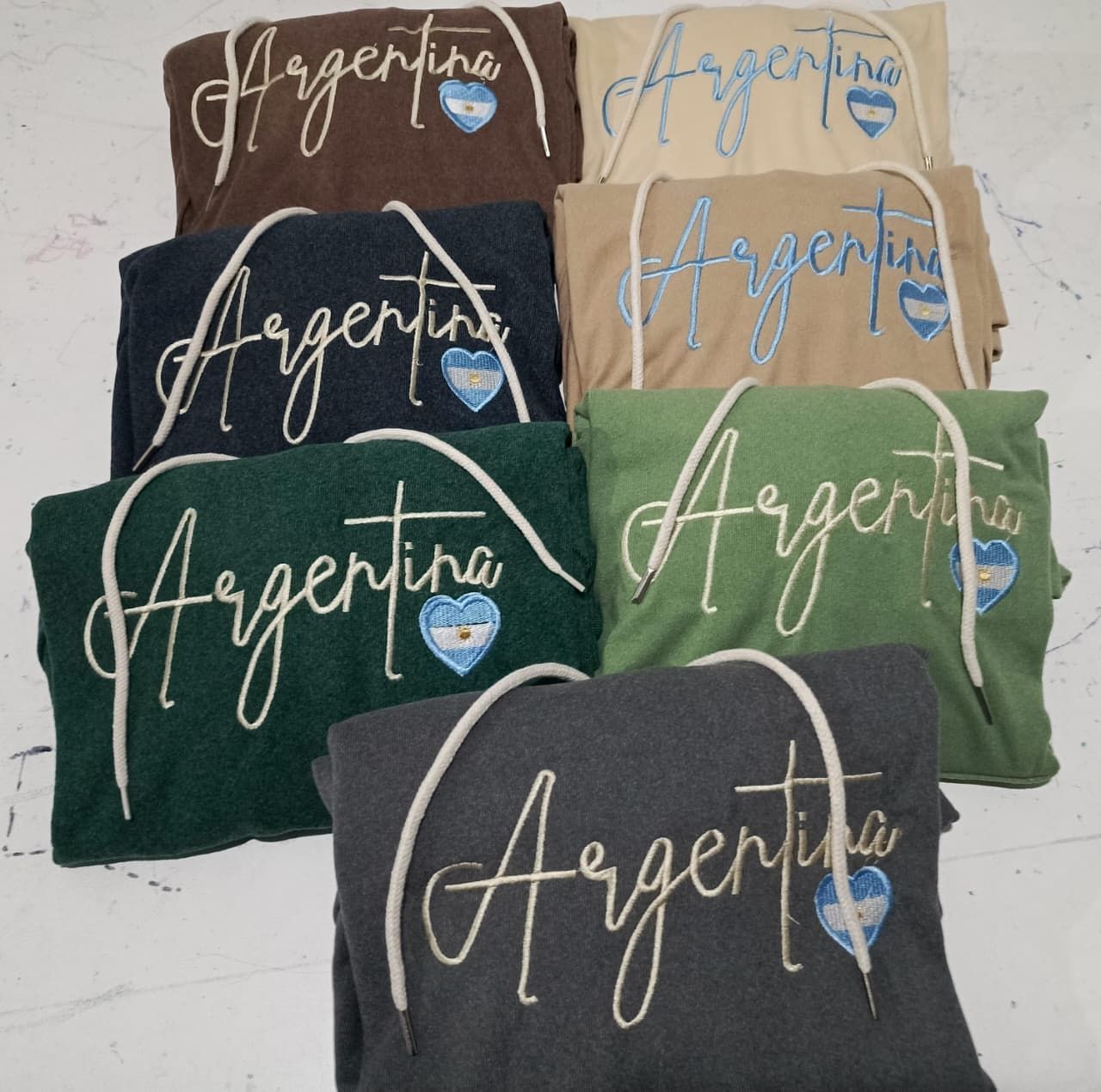 Sudaderas apiladas de Argentina en marrón, beige, negro, verde y gris con diseños de corazones azules y blancos.