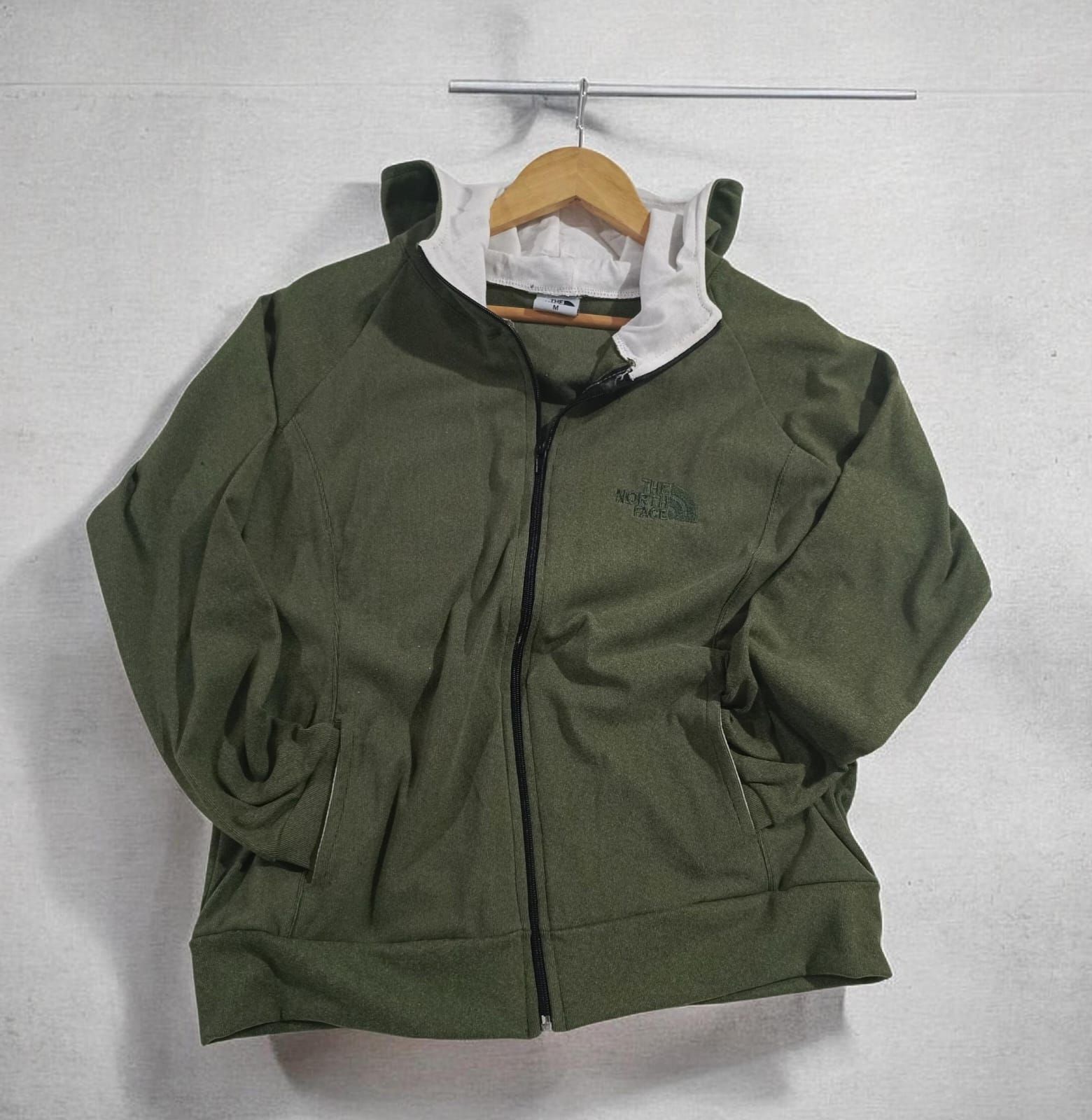 Chaqueta verde con cremallera colgada en una percha de madera contra una pared clara.