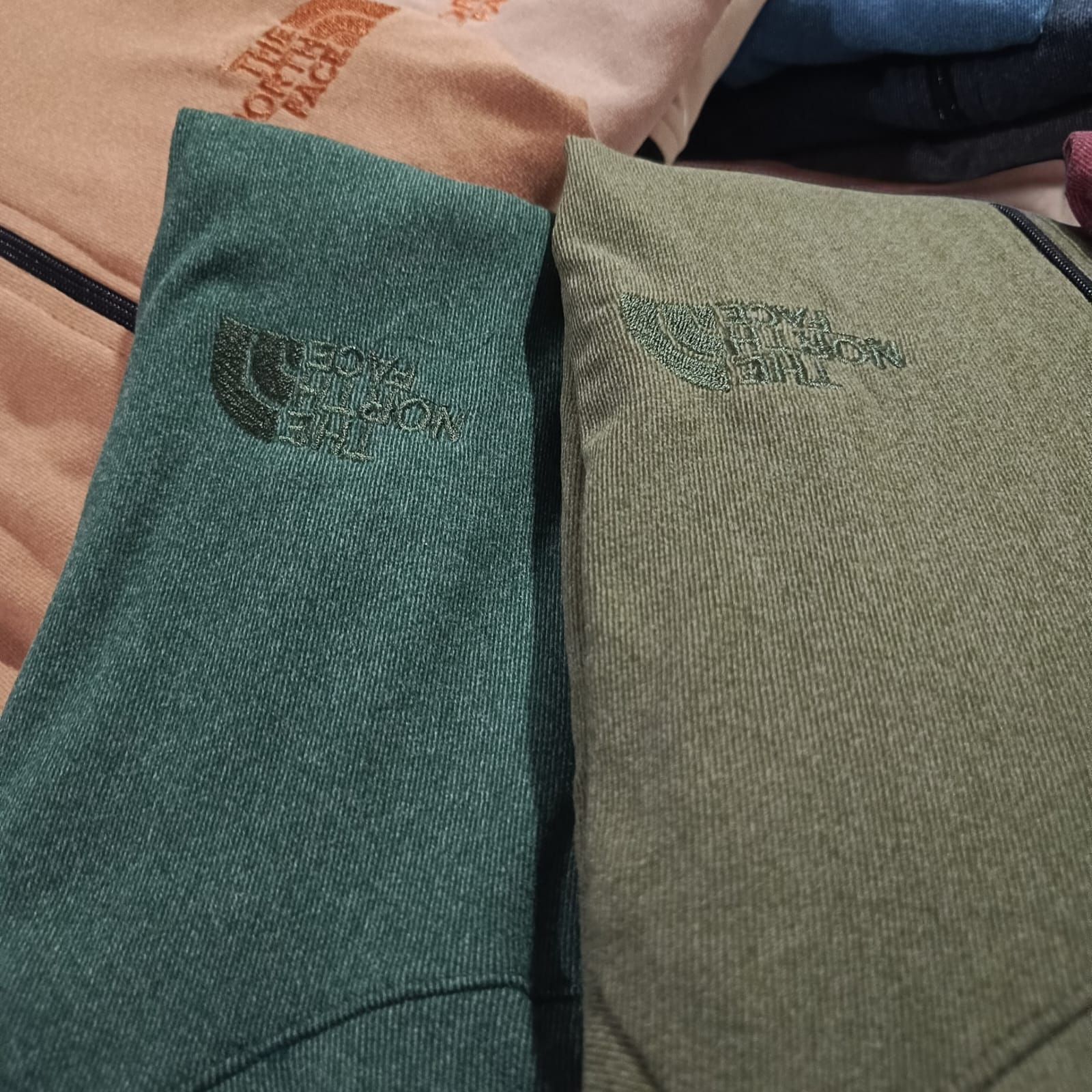 Chaqueta verde con cremallera colgada en una percha de madera contra una pared clara.