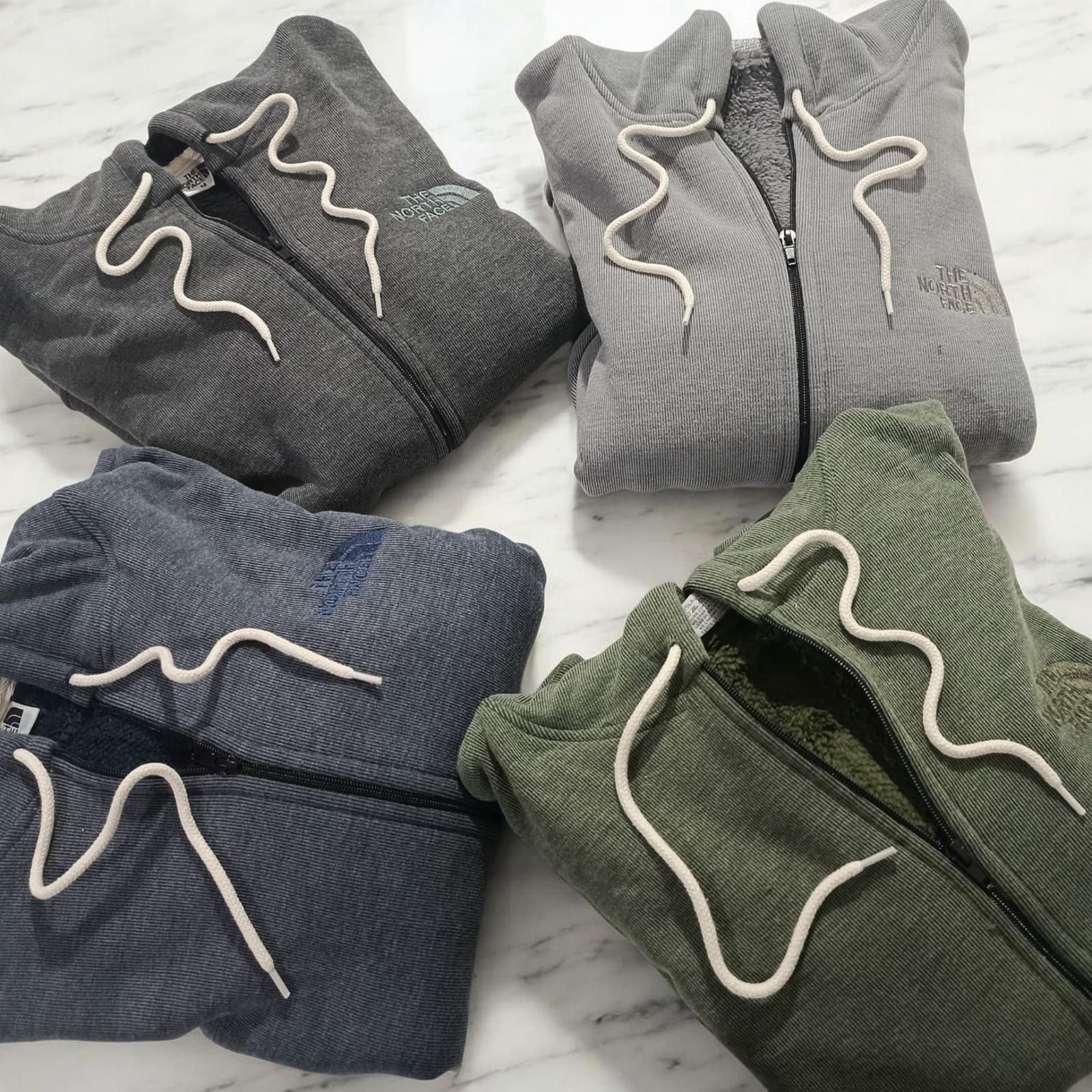 Sudaderas con capucha dobladas apiladas en gris oscuro, negro, gris claro y verde oliva sobre una superficie blanca.