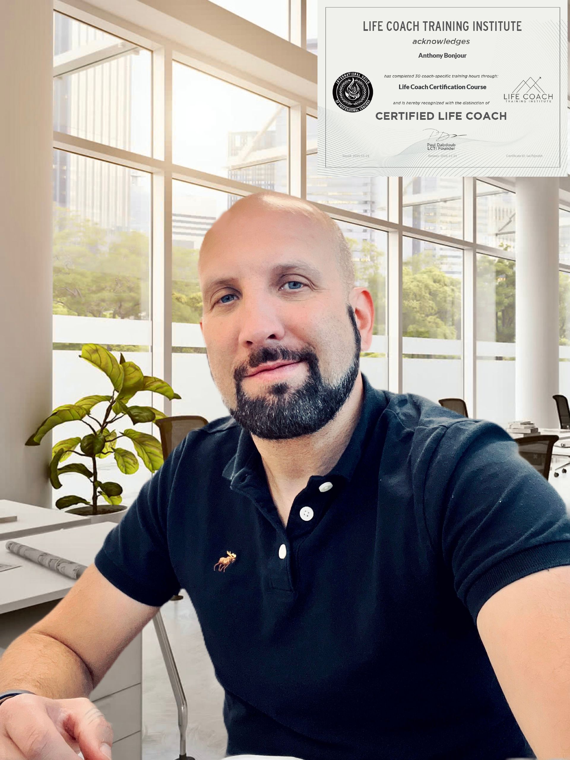 Man in black polo shirt at desk in bright office, looking at camera and smiling.