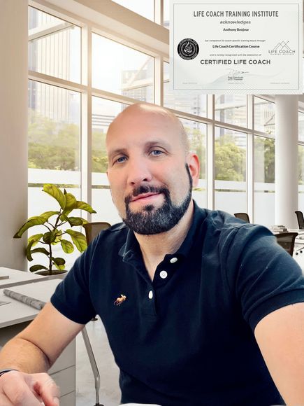 Man in black polo shirt at desk in bright office, looking at camera and smiling.