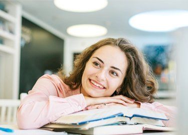 Woman with brown hair smiles, leaning on stacked books in a library, looking up with contentment.