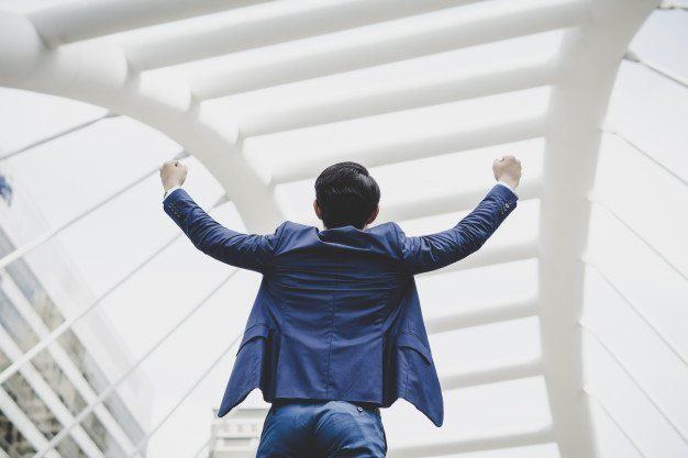 Man in a blue suit with arms raised in celebration stands under a white architectural structure.