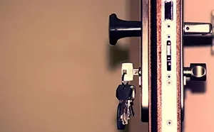 A close-up of a metal door lock mechanism with keys dangling from the keyhole against a plain, light-colored background.