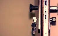 A close-up of a metal door lock mechanism with keys dangling from the keyhole against a plain, light-colored background.