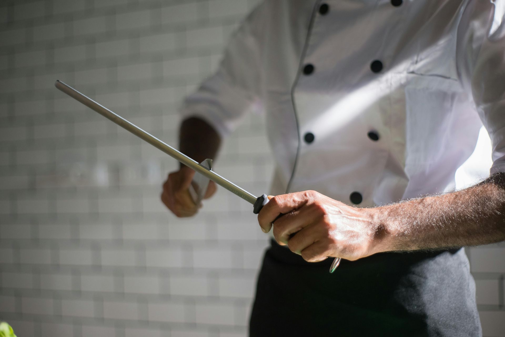 A chef in a white uniform uses a honing rod to sharpen a kitchen knife against a white brick wall background.
