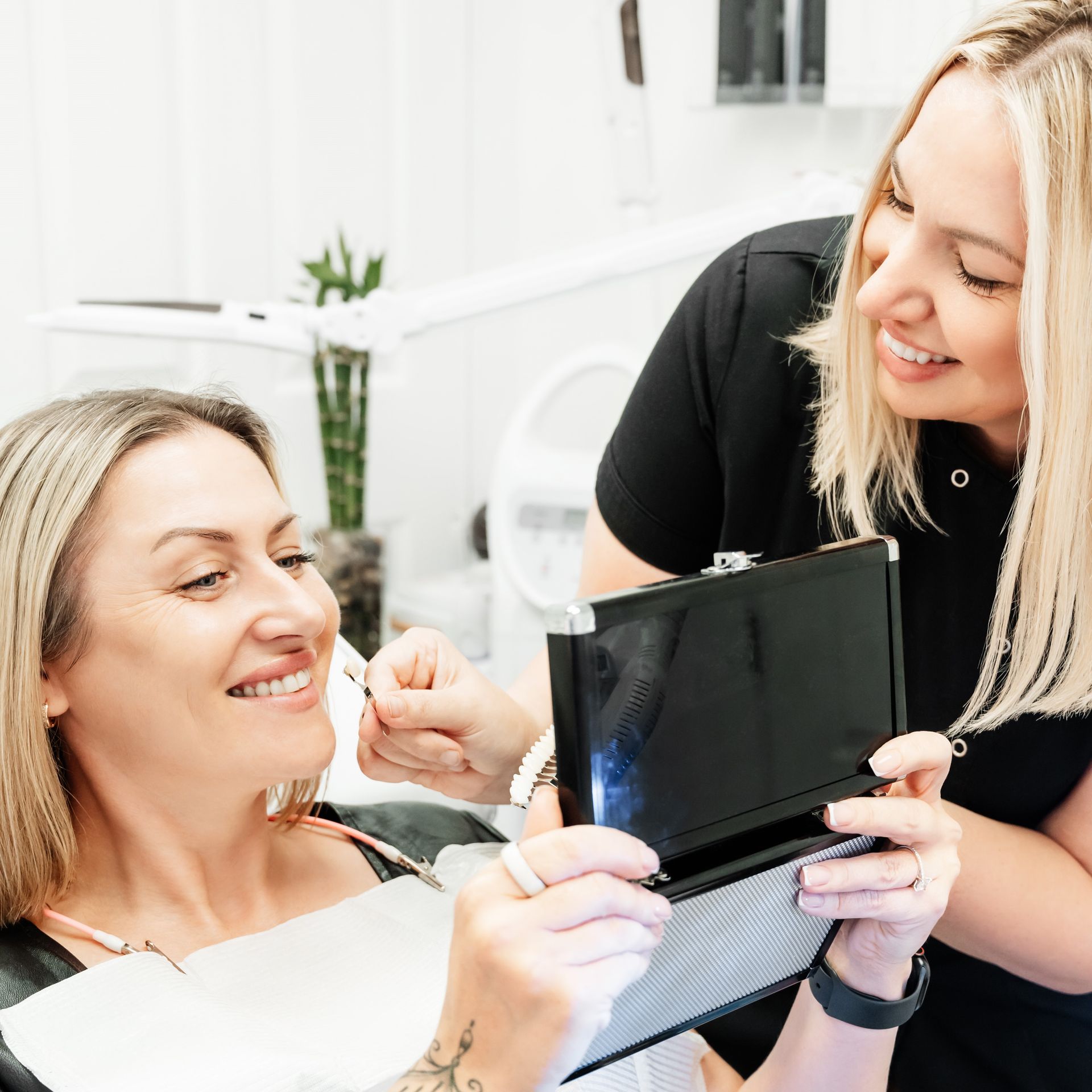 A woman is looking at her teeth in a mirror while another woman holds a tablet.