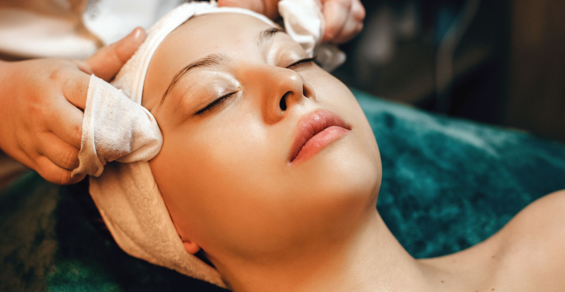 Woman receiving a facial treatment with closed eyes, head wrapped in a towel, towels on forehead.