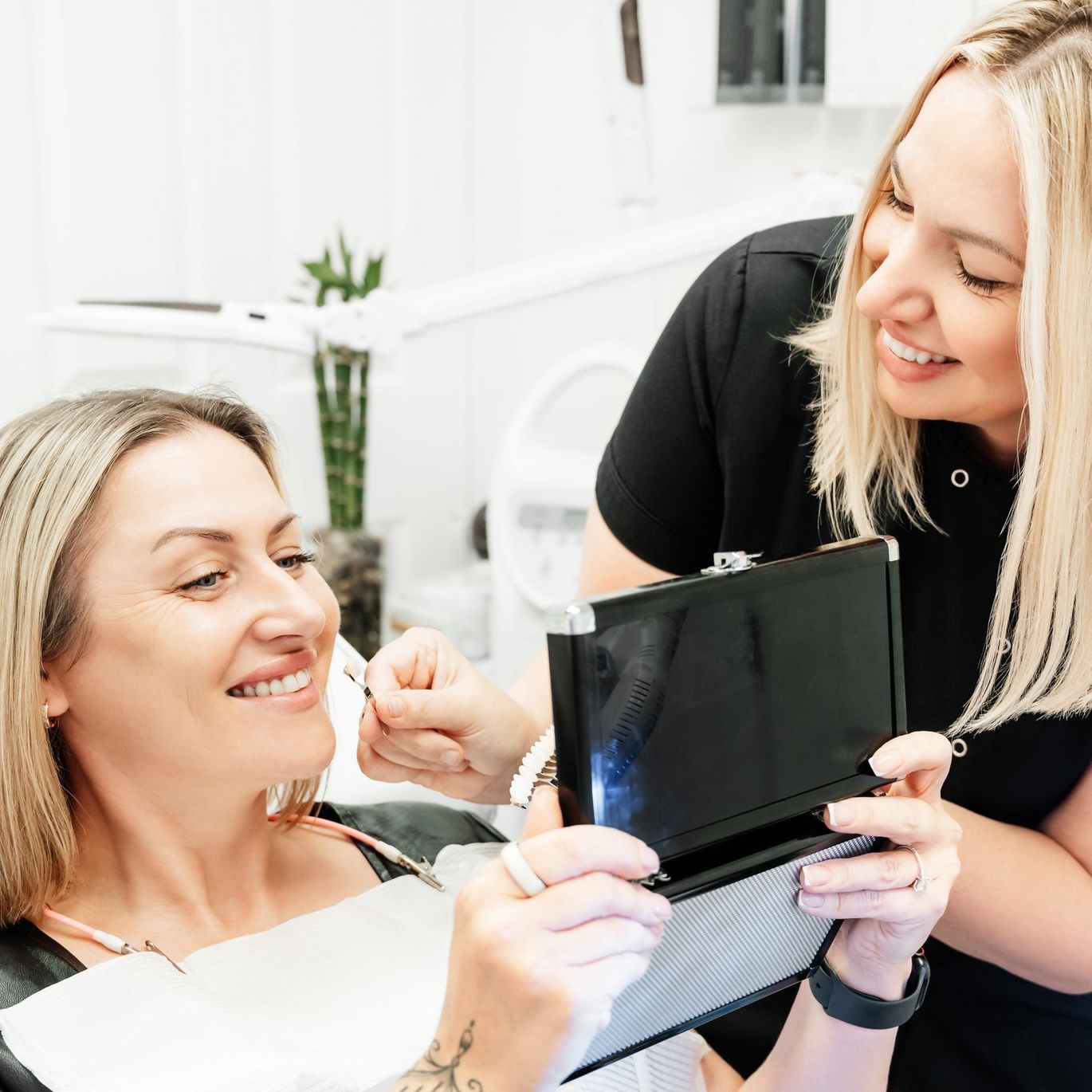 a woman is looking at her teeth in a mirror while another woman holds a tablet .