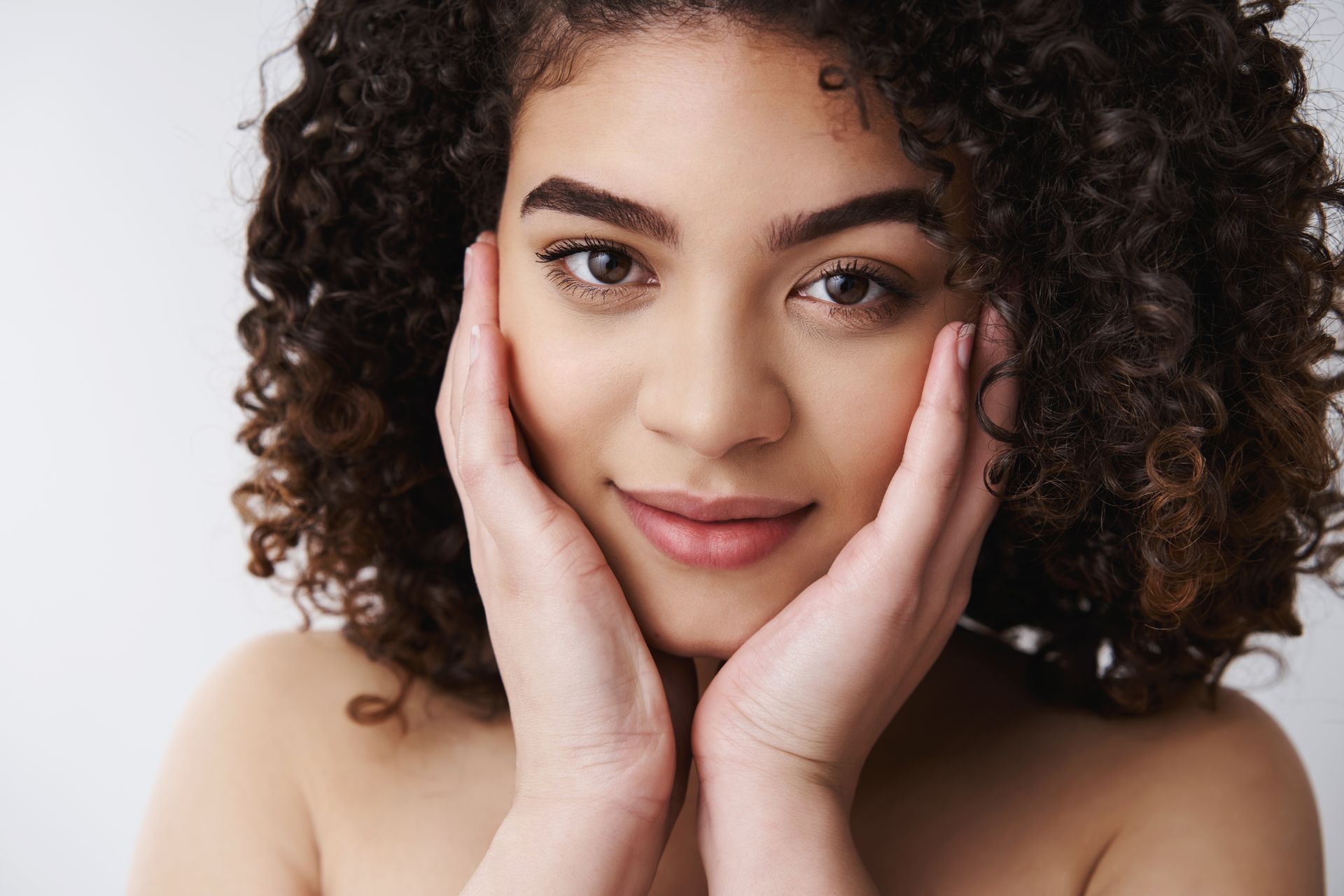 a woman with curly hair is touching her face with her hands and smiling .