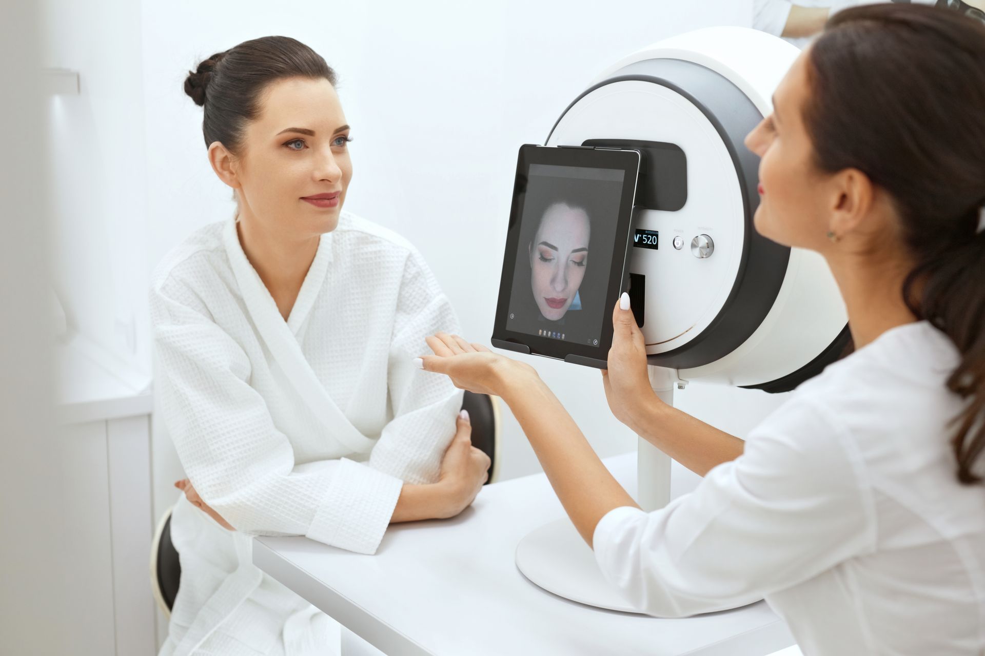 A woman is sitting at a table looking at a tablet with a picture of her face on it.