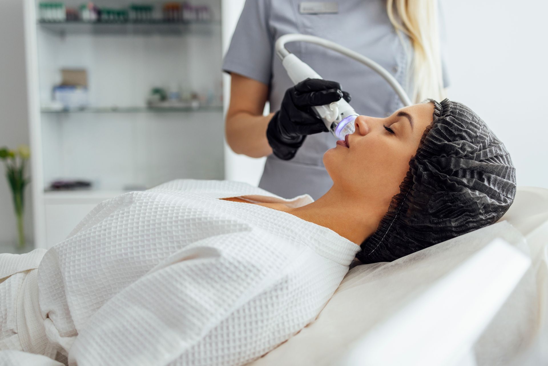 Woman receiving facial treatment in a clinic; a medical professional holds a device near her face.