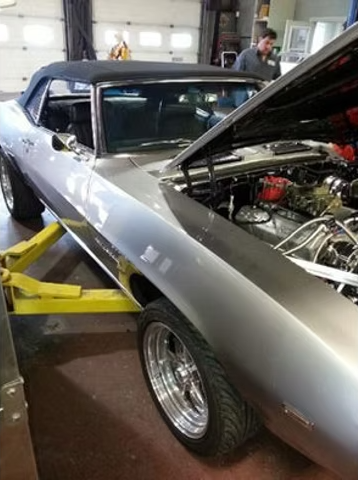 Silver convertible car with hood open in a repair shop, on a yellow lift. A mechanic is visible in the background. | Acadia Auto Service