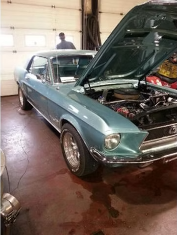 A teal 1968 Ford Mustang with the hood up, in a garage. A person stands in the background. | Acadia Auto Service