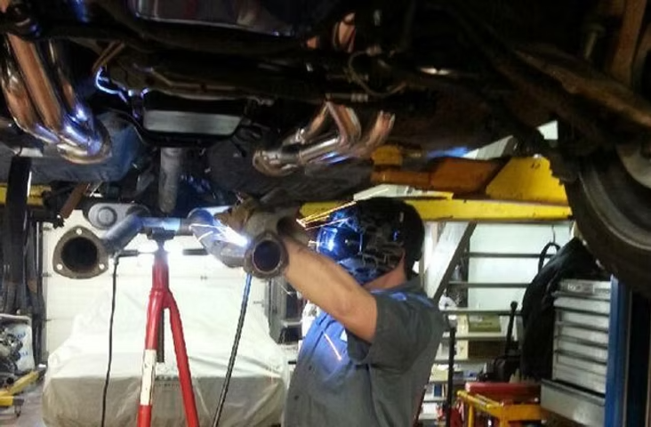 Mechanic welding exhaust system under a raised vehicle. Protective mask, shop setting. | Acadia Auto Service