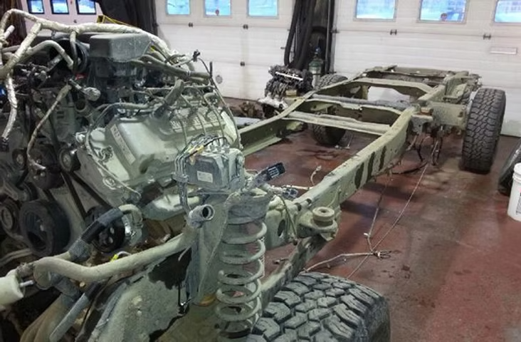 A disassembled truck chassis with engine and other parts in a garage, painted olive drab. | Acadia Auto Service